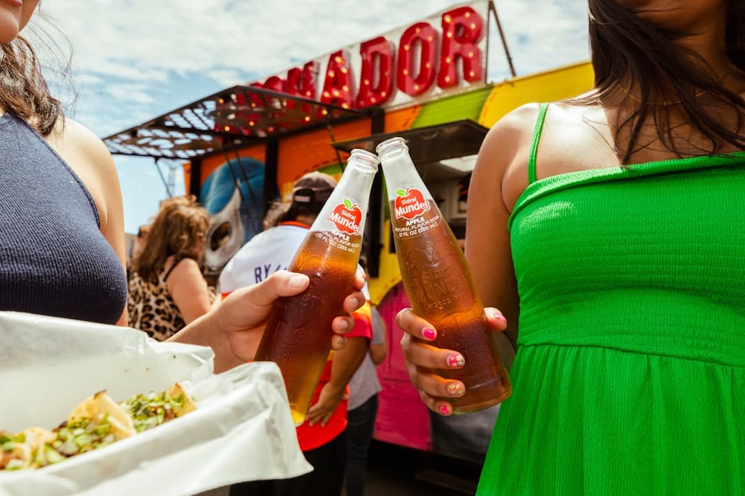 Two women toast with bottles of Mundet apple soda at a crowded outdoor fair; a colorful food stand is in the background, and a taco is visible in the foreground.