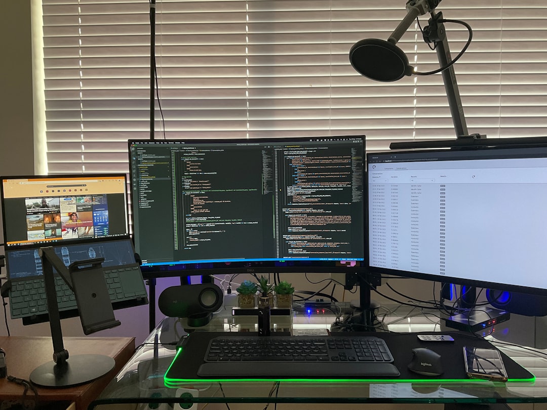 Triple-monitor coding workstation with a microphone, laptop on a stand, and a backlit glass desk setup.