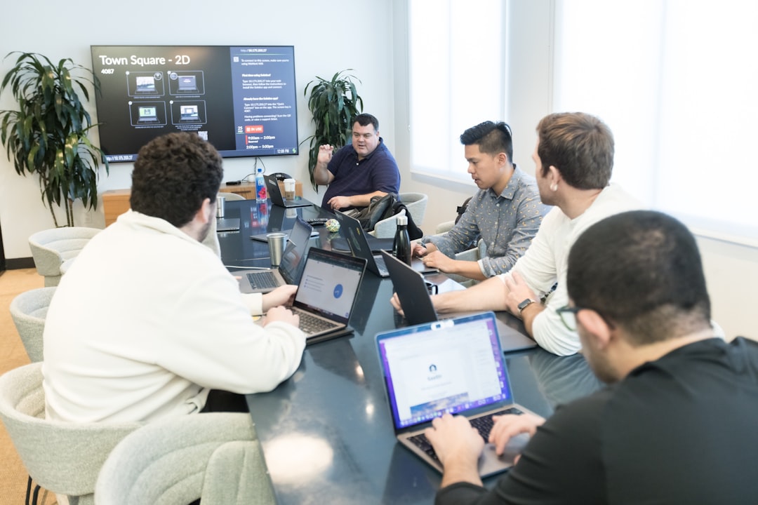 Team of six around a conference table with laptops; presenter at the head gesturing toward a screen labeled 'Town Square - 2D'.