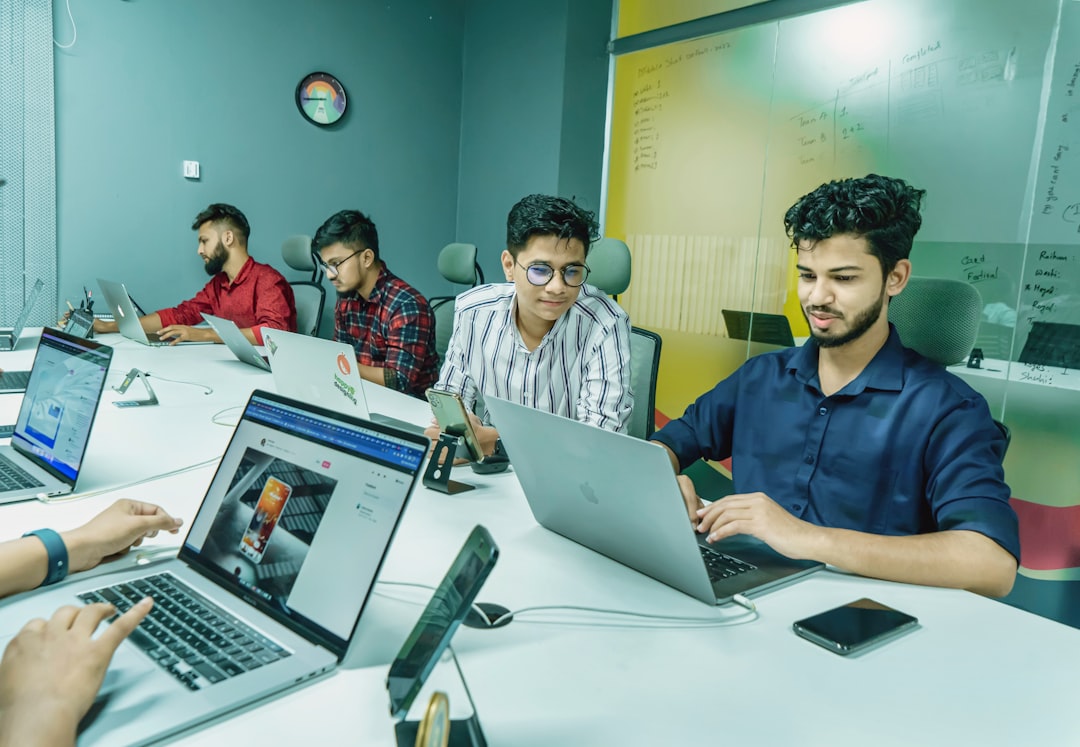 Group of professionals working on laptops around a conference table in a bright, glass-walled office.