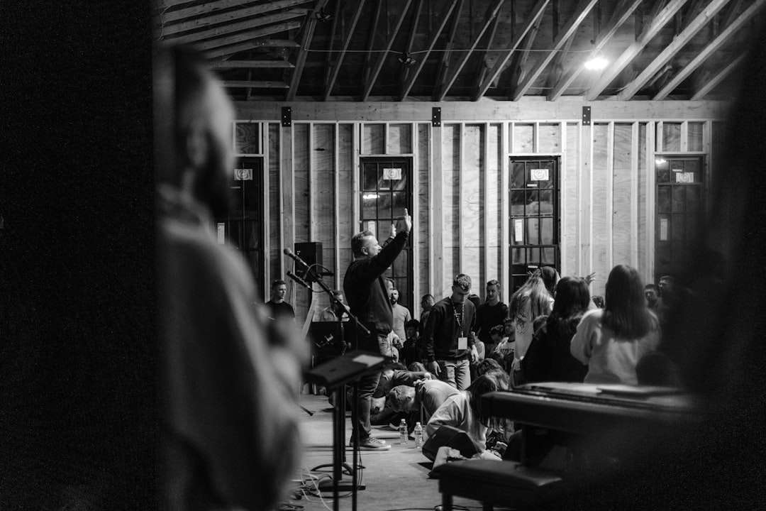 Man on stage with arms raised addressing a crowd in a bare-framed room, audience seated and standing around.
