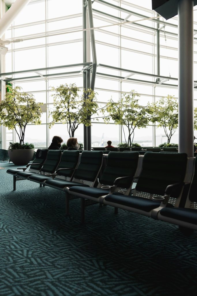 Row of green airport seating in a bright atrium with potted trees and large glass walls used for passengers waiting areas