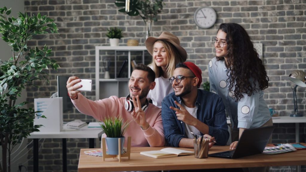 Four coworkers take a selfie in a brick-walled office, smiling at the camera with laptop and notebooks on the desk.
