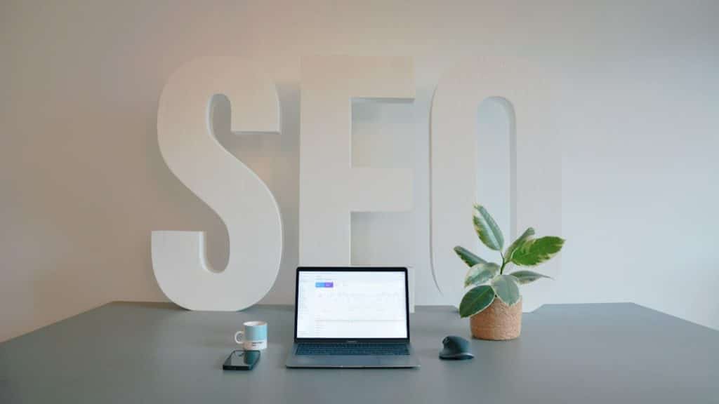 Desk setup featuring oversized white 'SEO' letters, a laptop with charts, a plant, a mug, phone and mouse on a gray desk.