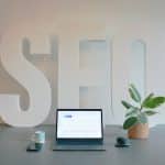 Desk setup featuring oversized white 'SEO' letters, a laptop with charts, a plant, a mug, phone and mouse on a gray desk.