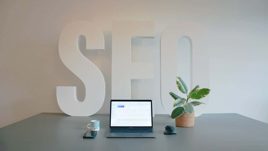 Desk setup featuring oversized white 'SEO' letters, a laptop with charts, a plant, a mug, phone and mouse on a gray desk.