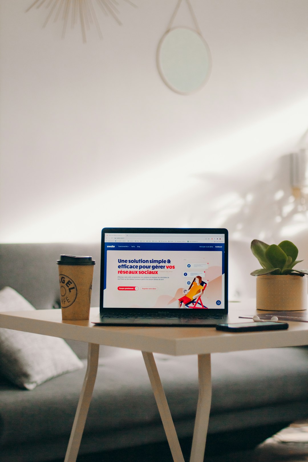 Laptop on a white table displaying a colorful website; a coffee cup sits to the left and a small plant to the right in a bright room.