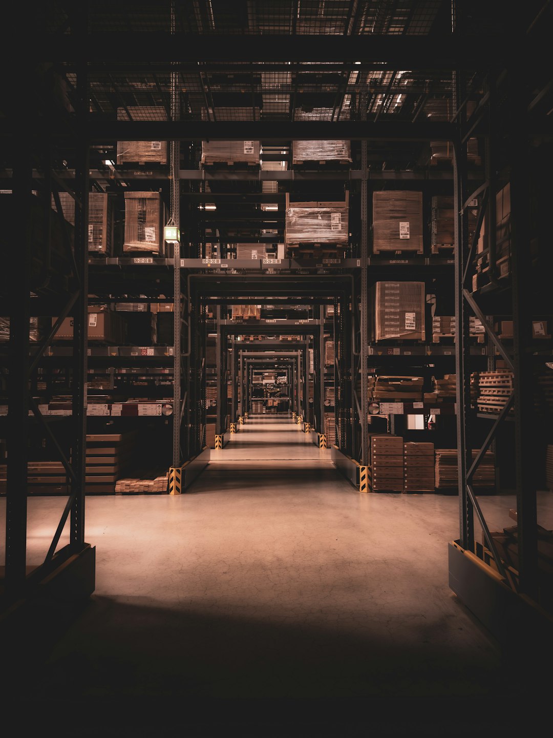 Dimly lit warehouse aisle with tall metal racks and stacked pallets receding into the distance, creating a tunnel-like perspective.