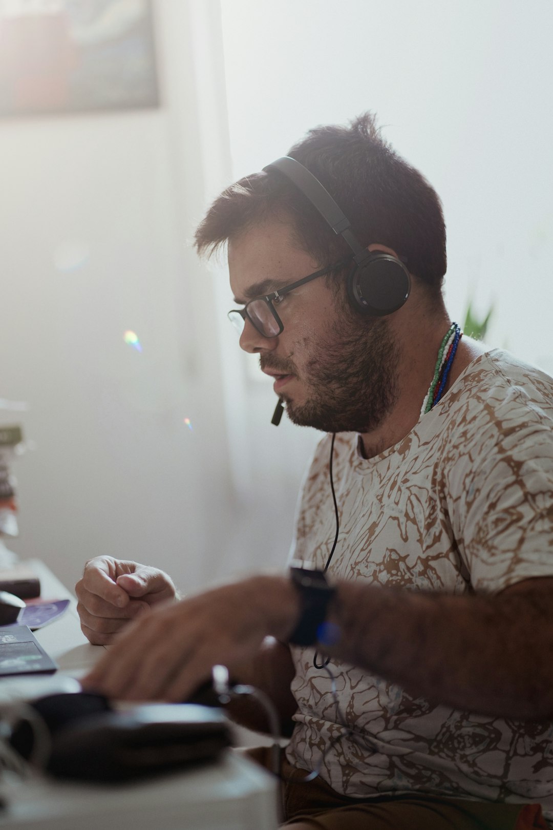 Man wearing over-ear headphones and a microphone headset working at a desk, looking at a computer screen in a bright room.