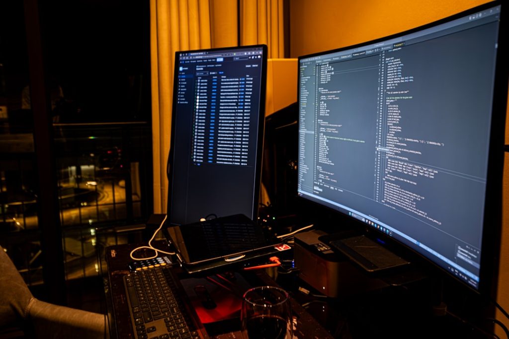 Dimly lit workstation with two monitors displaying code, a laptop, keyboard, and a glass on the desk.