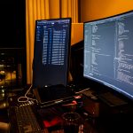 Dimly lit workstation with two monitors displaying code, a laptop, keyboard, and a glass on the desk.