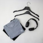 Folded gray t-shirt with a small logo on top of a black shirt, plus a pair of over-ear headphones and two empty black hangers on a light background.