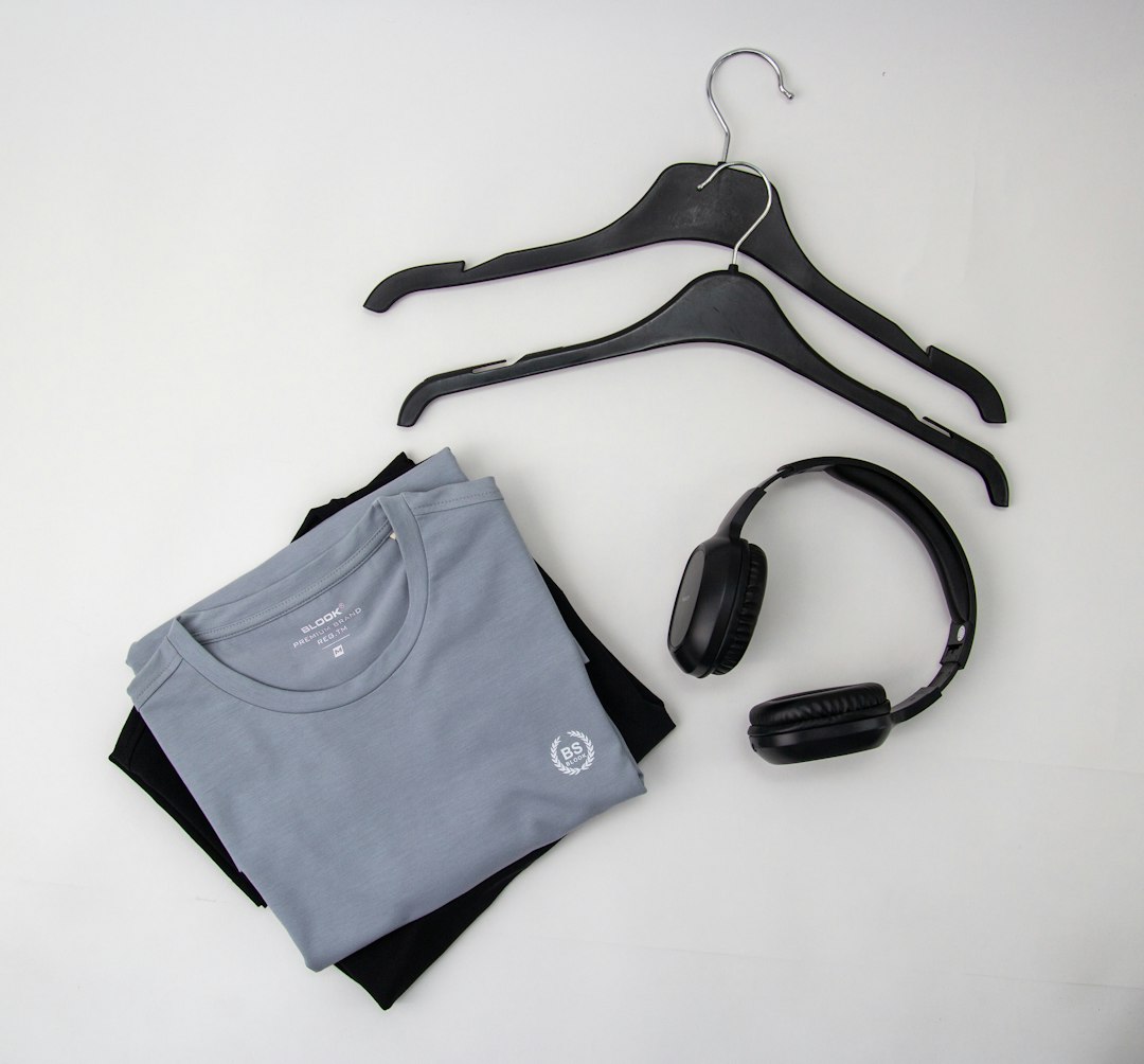 Folded gray t-shirt with a small logo on top of a black shirt, plus a pair of over-ear headphones and two empty black hangers on a light background.