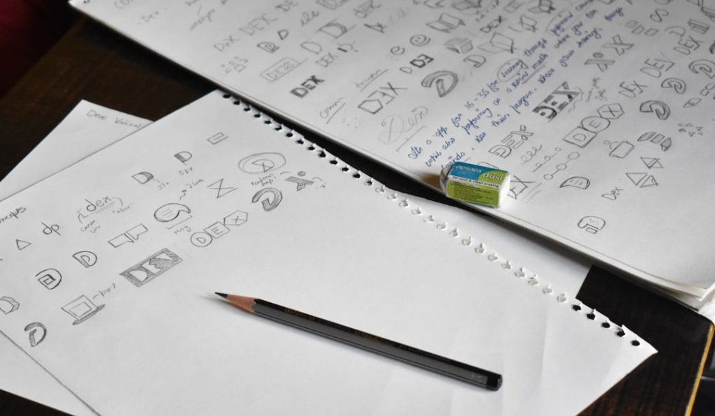 Sketchbook pages with hand-drawn icons and symbols (like folders, shapes, and designer logos) scattered across a desk next to a pencil and an eraser on spiral-bound paper