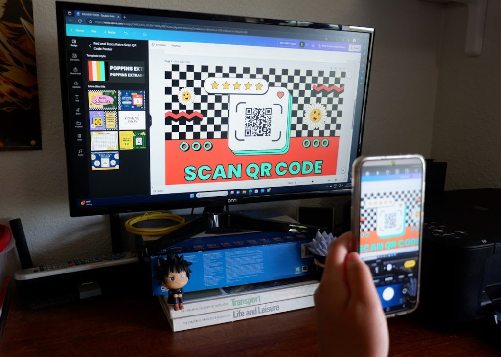Monitor showing a colorful poster that reads SCAN QR CODE with a QR code; a hand holds a smartphone taking a photo of it.