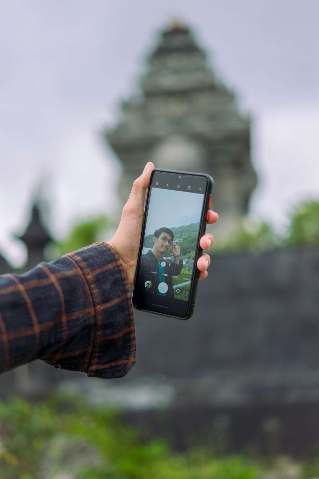 Person holding a smartphone to take a selfie, with a temple and greenery blurred in the background.