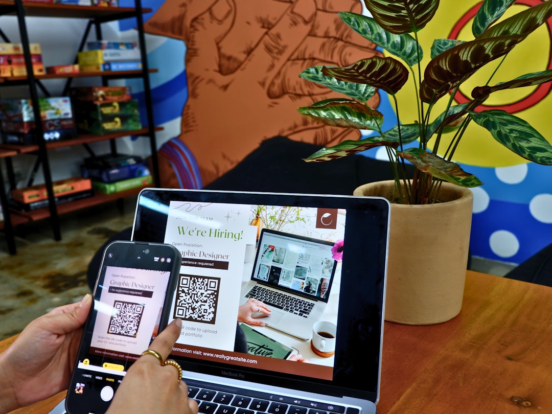 Person holds a smartphone to scan a QR code on a 'We're Hiring!' poster shown on a laptop screen, with a plant and colorful background nearby.