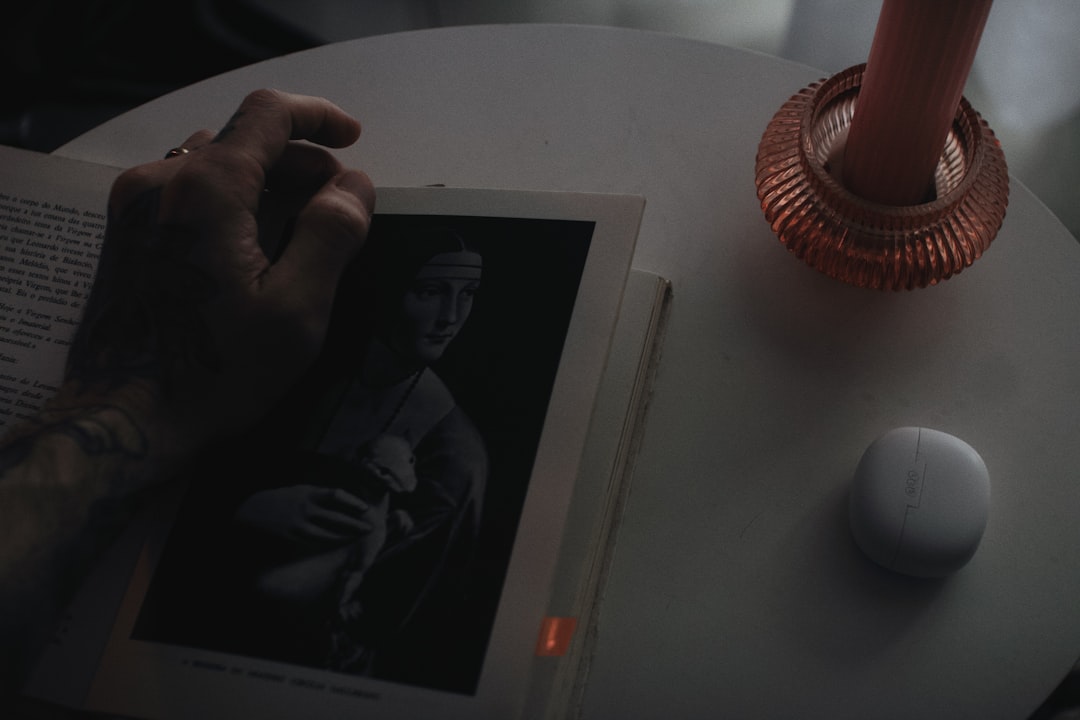 Tattooed hand turning a page in a book opened to a monochrome portrait of Mona Lisa on a round table with a pink ribbed vase and a small white gadget.