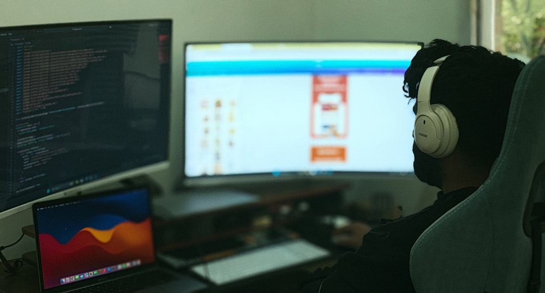 Person wearing white over-ear headphones sits at a desk with two monitors and a laptop, coding on the left screen.