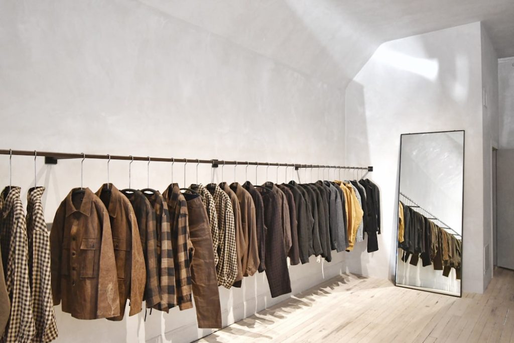 Clothes rack with brown and checkered jackets in a minimalist store; a full-length mirror leans against the wall to the right.