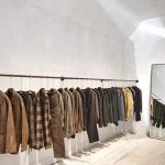 Clothes rack with brown and checkered jackets in a minimalist store; a full-length mirror leans against the wall to the right.