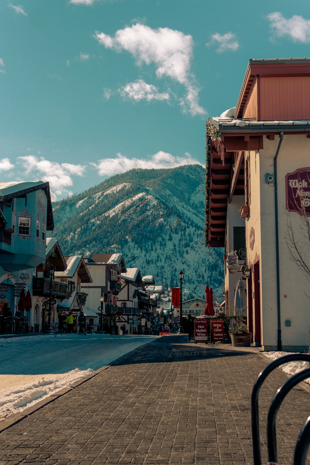 Snowy mountain street lined with colorful shops, blue sky, with pine-covered hills in the background.