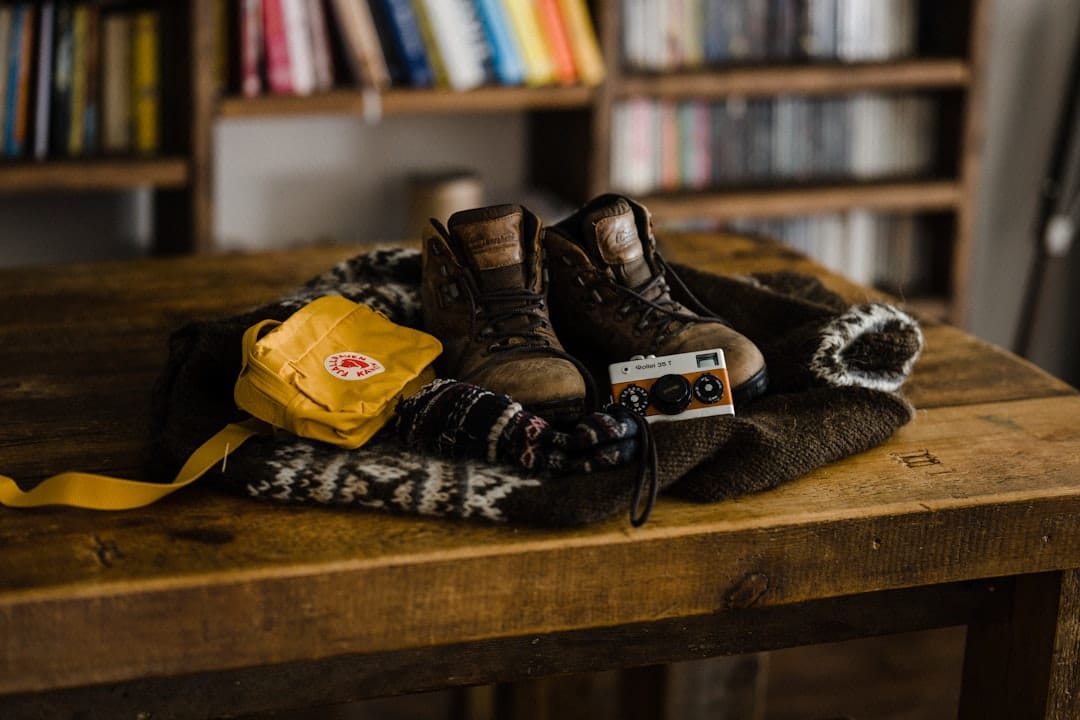Brown hiking boots sit on a wooden table beside a patterned scarf, a yellow bag, and a small vintage camera lined up in a cozy room.