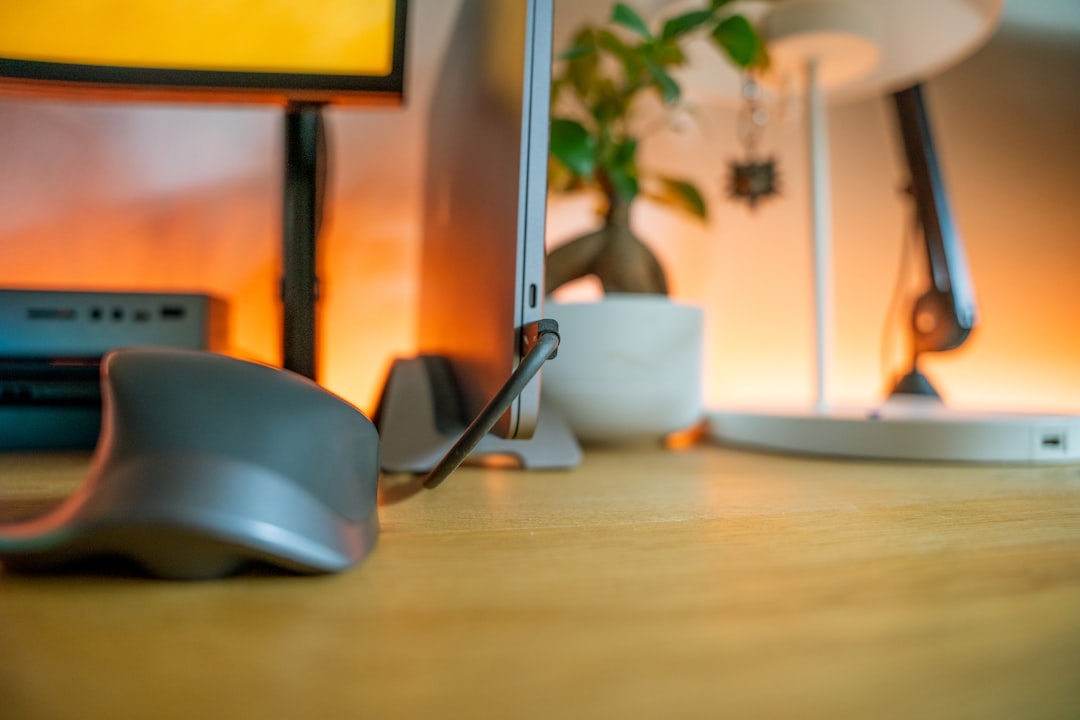 Desk setup: gray computer mouse in the foreground, monitor to the left, plant and arc lamp in warm background lighting.