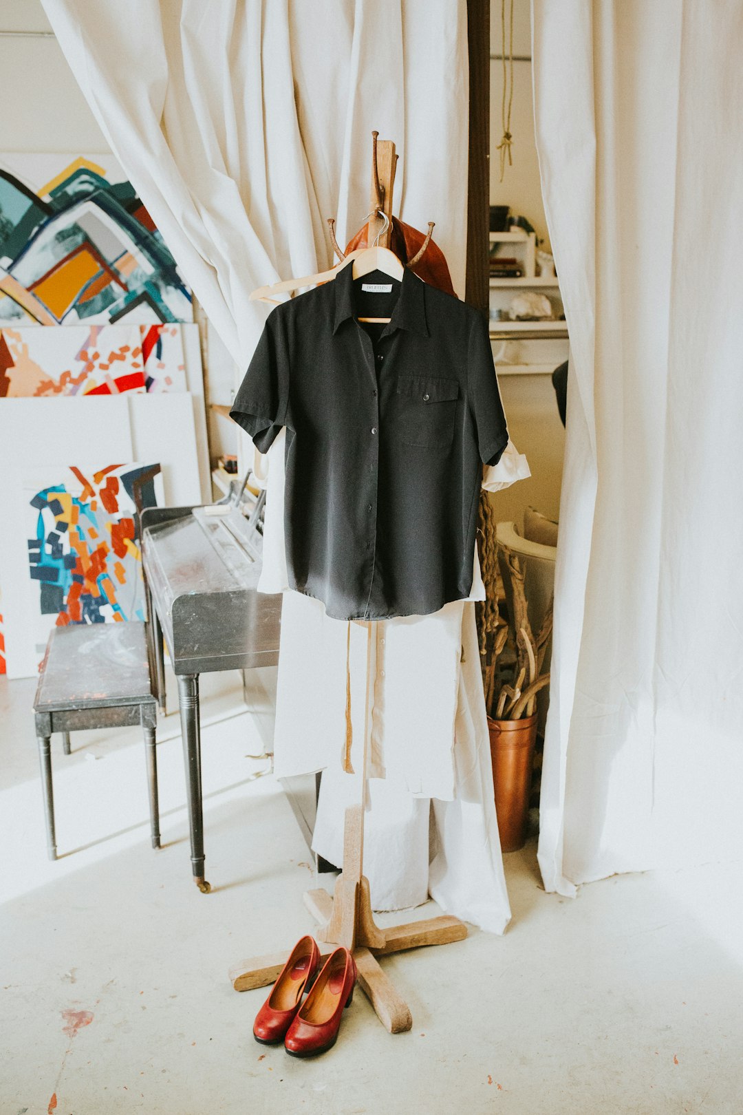 Black short-sleeve button-down shirt on a hanger in a bright studio with abstract paintings and white curtains; red shoes on the floor nearby.