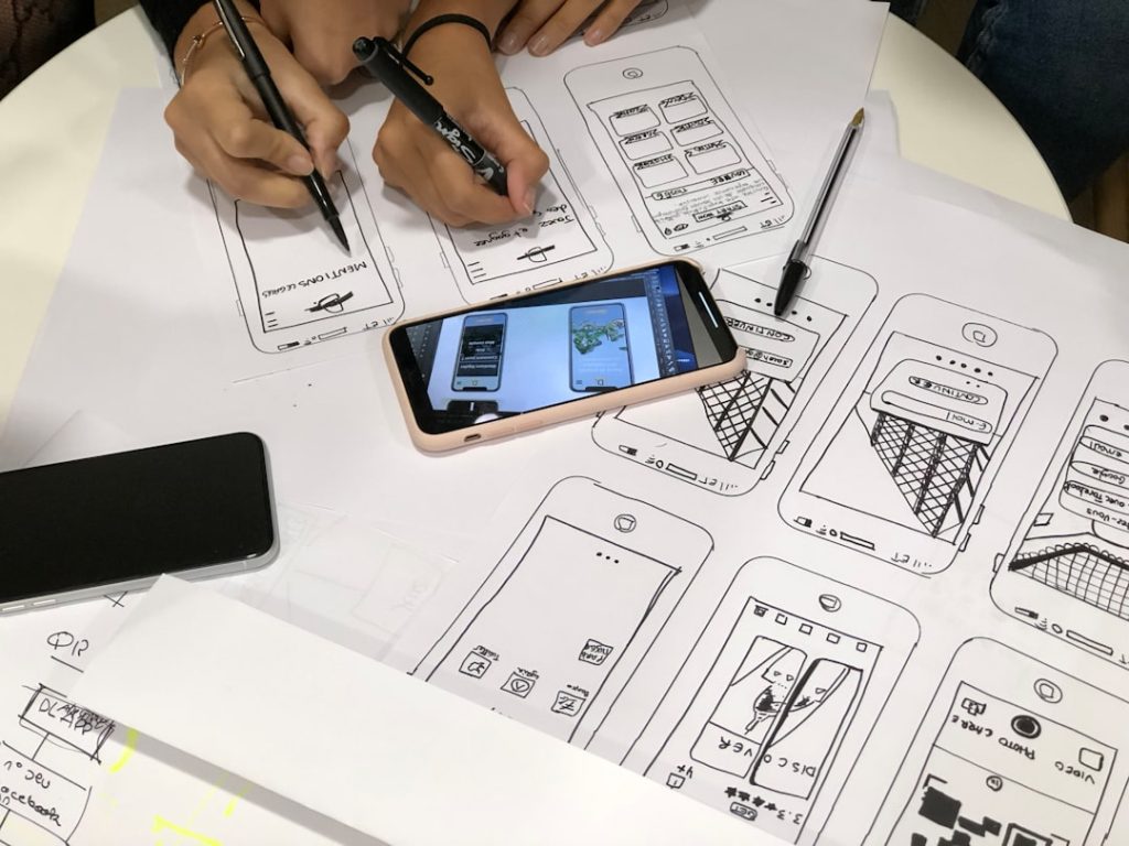 Design team sketching mobile app wireframes on large sheets, markers in hand, phones resting on the paper.