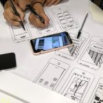Design team sketching mobile app wireframes on large sheets, markers in hand, phones resting on the paper.