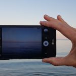 Hand holding a smartphone horizontally to photograph a sunset over calm ocean.
