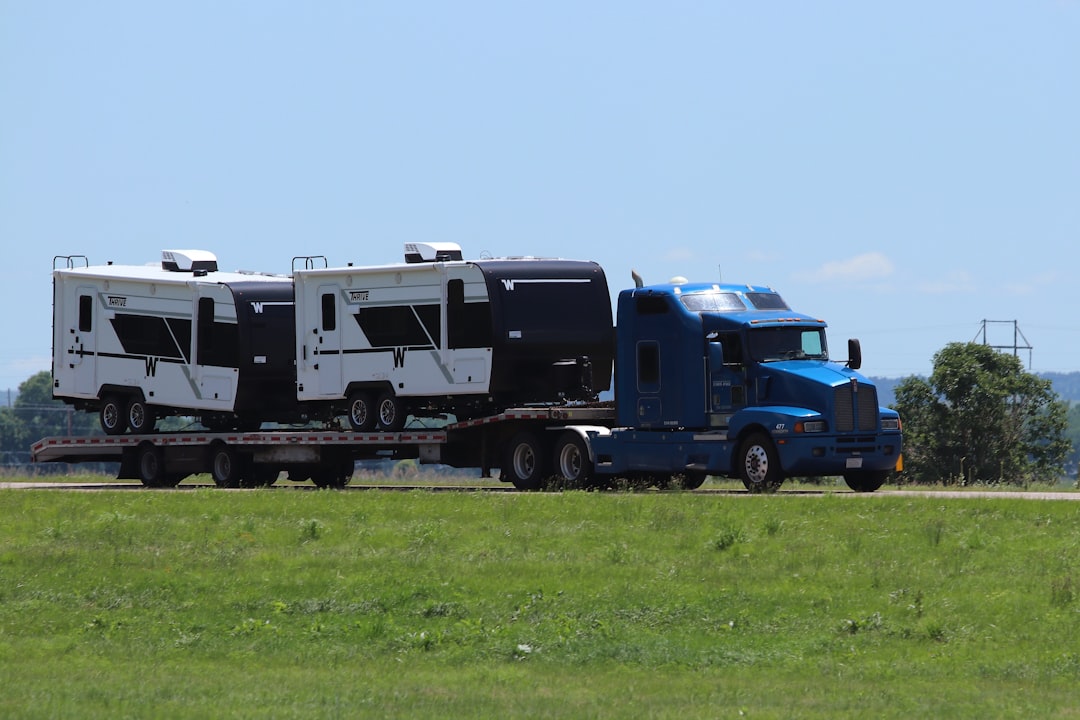 Blue semi-truck hauling a flatbed trailer loaded with three white travel trailers on a rural road under a clear sky.