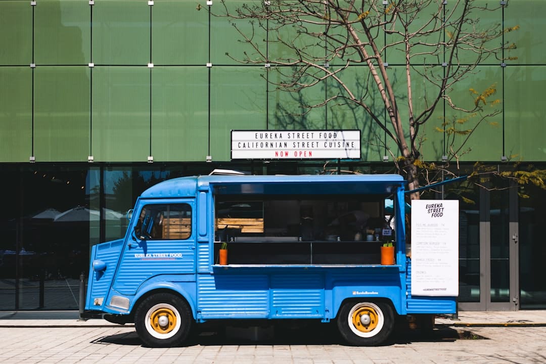 Blue vintage street-food truck parked along a sidewalk in front of a green paneled building, with a menu board on the side and small orange plant pots on the counter