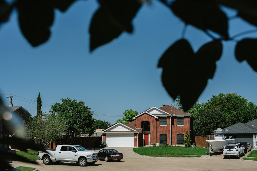 Leaves frame a sunny suburban scene: a brick two-story house with a white garage, green lawn, and several vehicles (white pickup, black car) plus a boat on a trailer in the driveway/side yard.