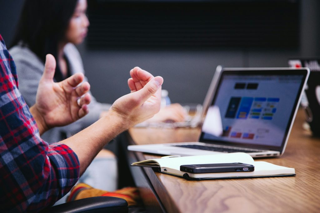 Hands gesturing in a meeting, with a laptop and notebook on a wooden table in the foreground.