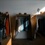 Clothes racks with jackets and sweaters in a minimalist shop, sunlight casting a diagonal shadow on the wall.