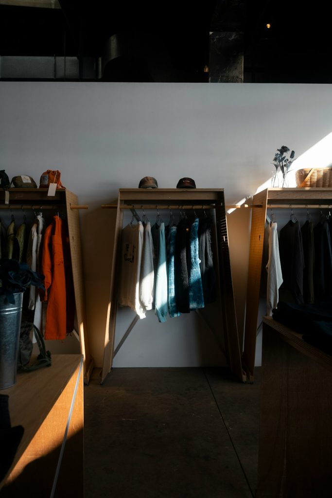 Clothes racks with jackets and sweaters in a minimalist shop, sunlight casting a diagonal shadow on the wall.