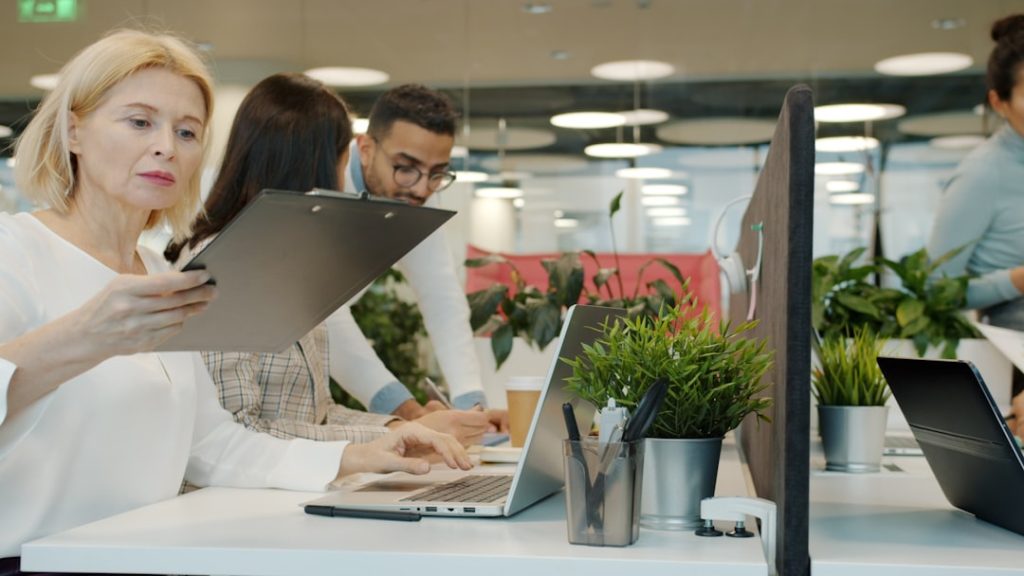 Woman holding a clipboard while two colleagues work on laptops at a bright office desk with plants nearby.