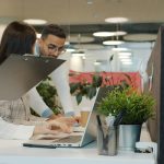 Woman holding a clipboard while two colleagues work on laptops at a bright office desk with plants nearby.