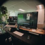 Video editing workstation with a large monitor showing a timeline, laptop, keyboard, mouse, headphones, and a leafy plant in a bright office.