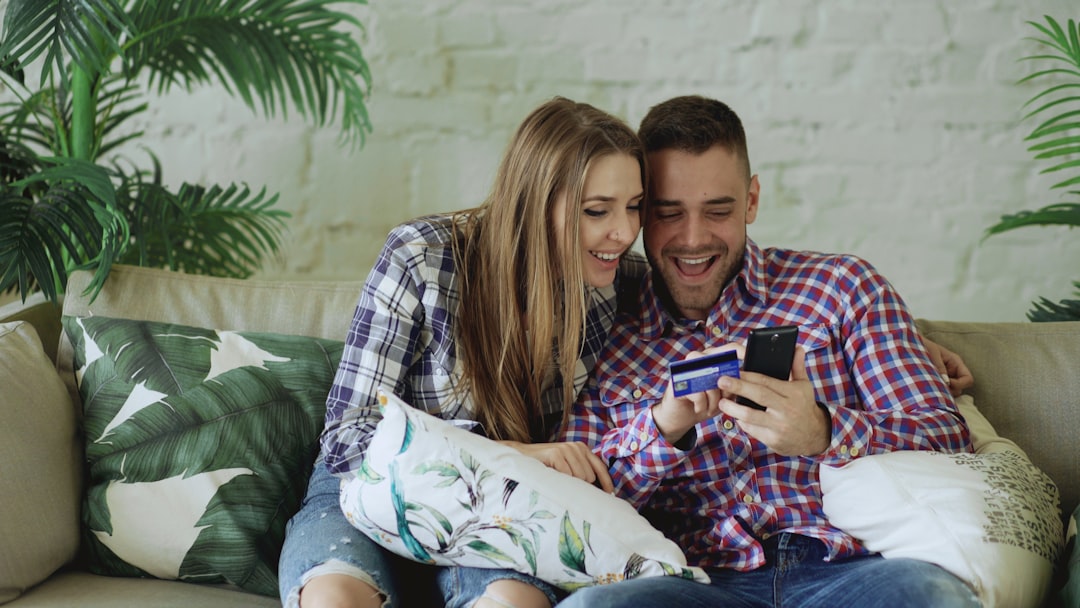 Couple sitting on a sofa, smiling as they shop online using a smartphone and credit card.