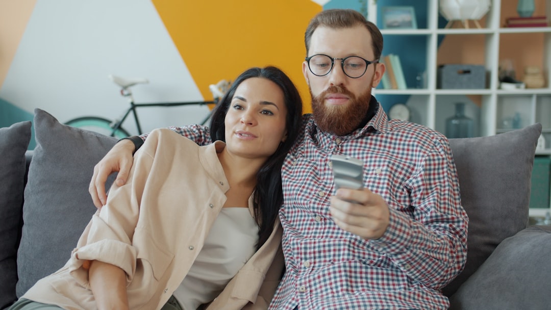 Couple sits close on a gray couch; the bearded man with glasses looks at a phone while the woman rests beside him in a colorful living room.