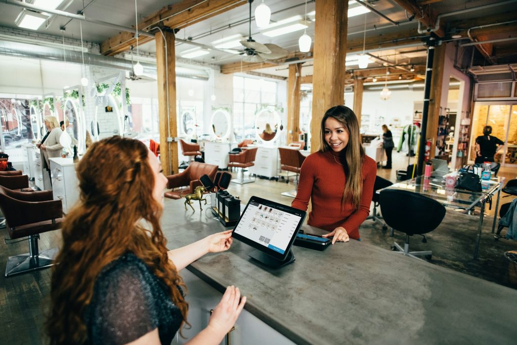Two customers at a salon reception counter using a tablet checkout system in a bright, modern space with mirrors and chairs in the background.