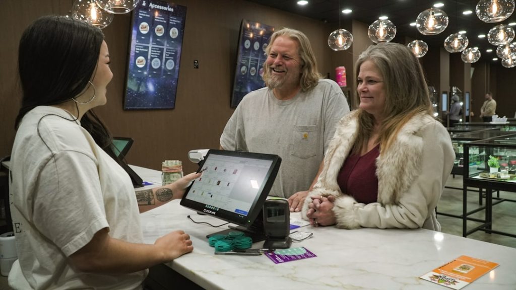 Cashier with a tattooed forearm operates a touchscreen POS for a smiling couple at a marble checkout counter in a store.