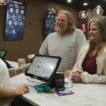 Cashier with a tattooed forearm operates a touchscreen POS for a smiling couple at a marble checkout counter in a store.