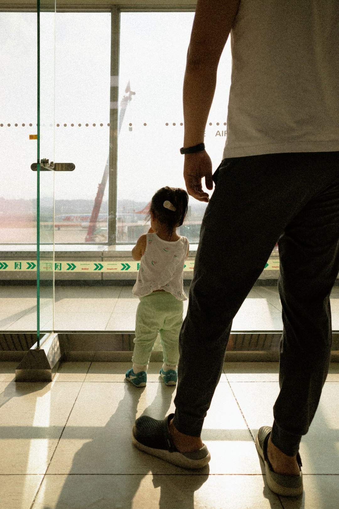 A toddler stands at an airport window, watching airplanes, with an adult man standing nearby in the foreground.