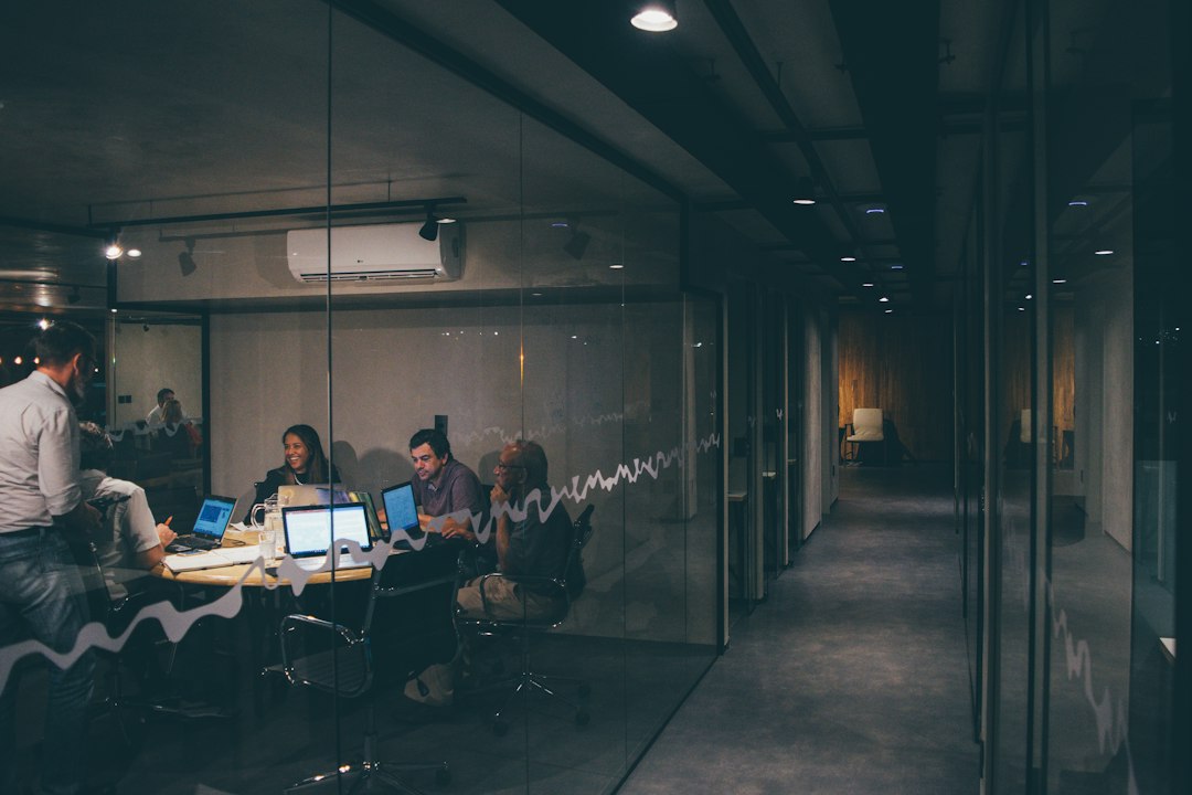 Team of professionals seated around a glass-walled conference room table with laptops, discussing work.