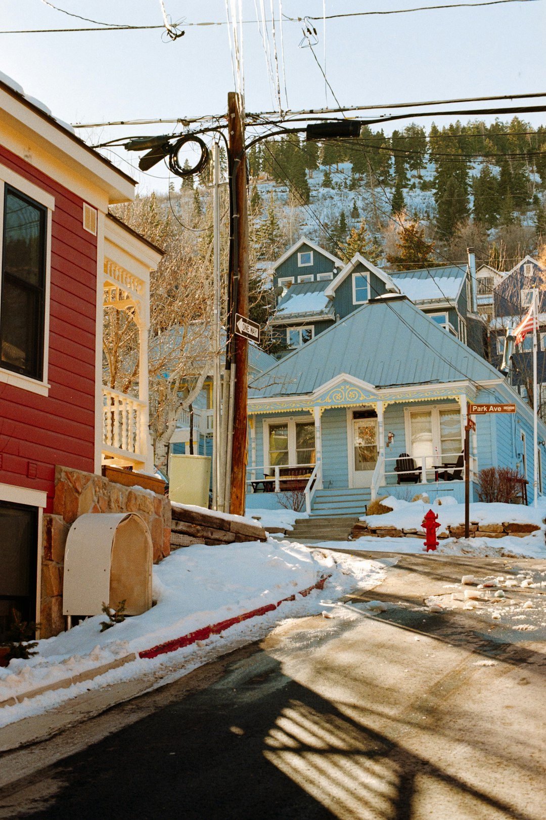 Snowy street in a mountain town with a red house foreground, blue house center, and tangled utility wires overhead on a sunny winter day.