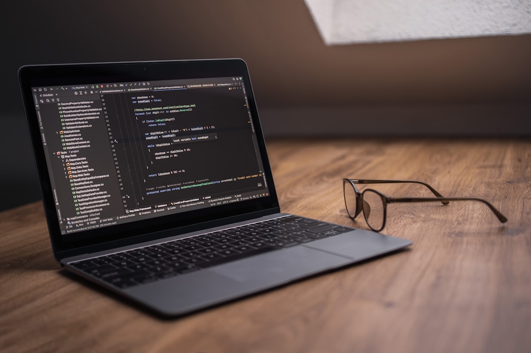 Laptop on a wooden desk displaying dark-mode code editor with lines of code, and a pair of glasses resting nearby on the right.